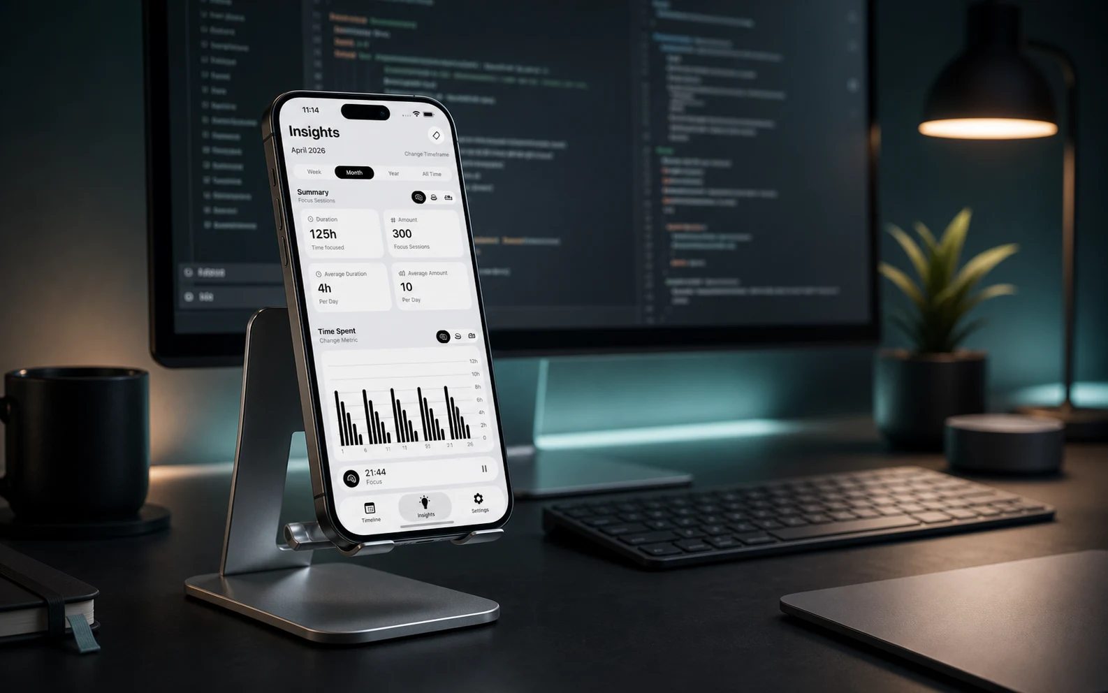 A smartphone on an aluminum desk stand showing FocusKit insights in front of a large monitor, keyboard, plant, and warm desk lamp.